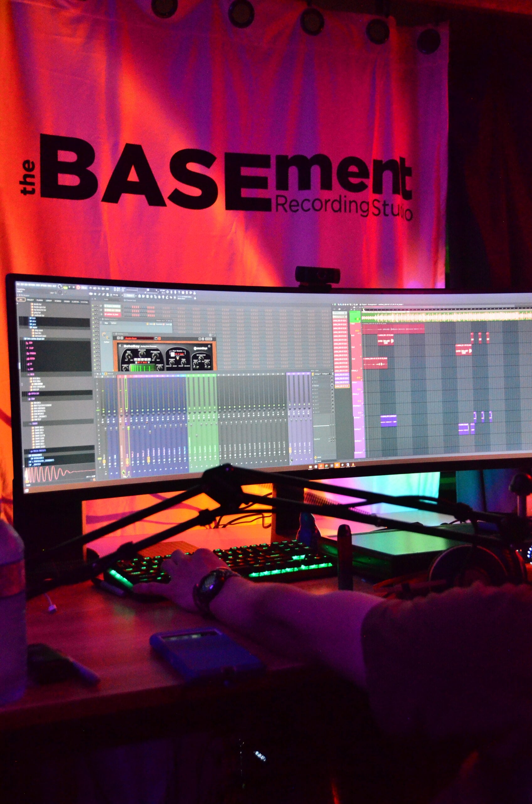 An ultrawide curved monitor showing a DAW mixer with dozens of channel faders and an arrangement window, the studio banner lit red behind it and an engineer typing on the desk